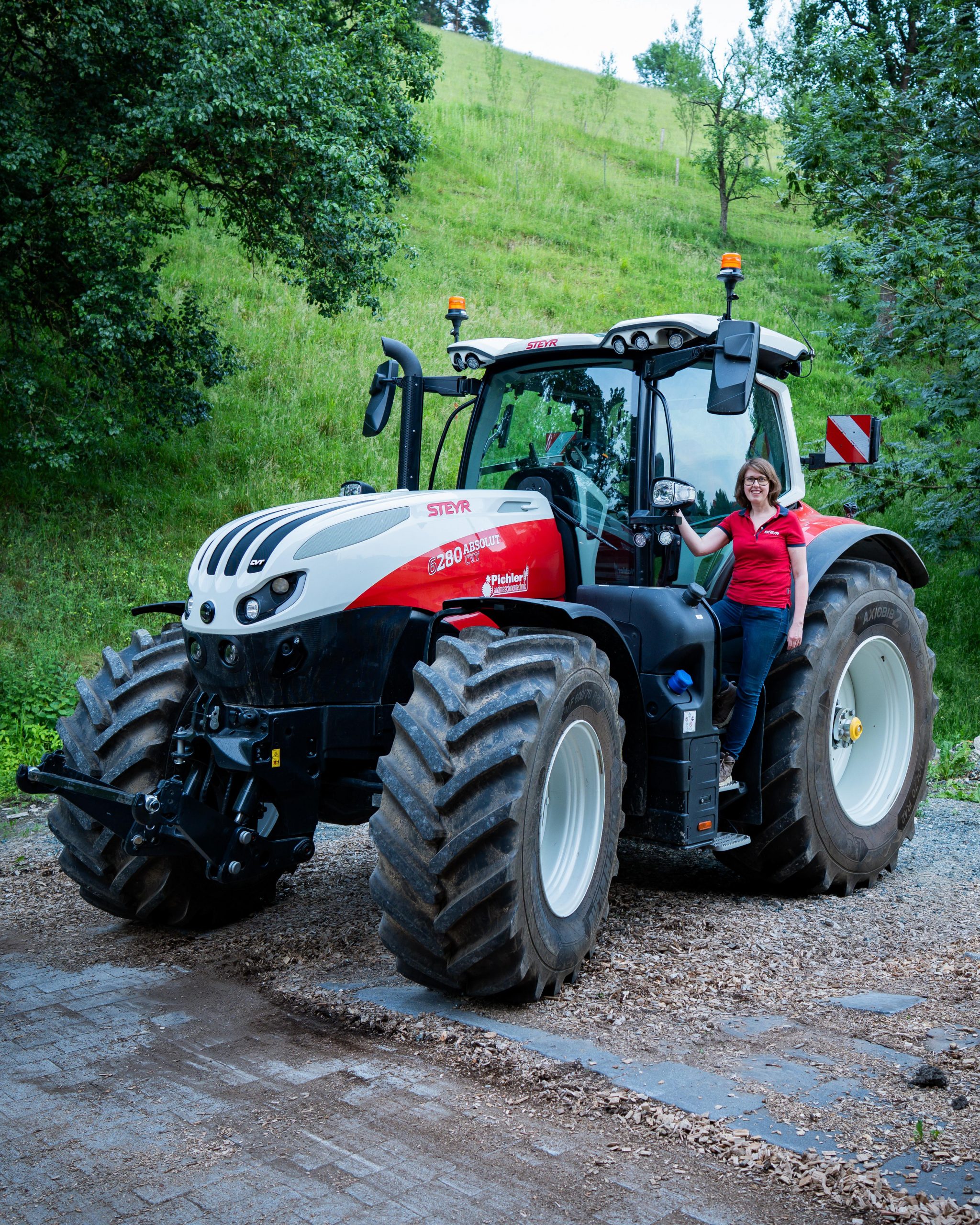 Ein moderner Steyr 6280 Absolut CVT Traktor mit großen Reifen steht auf einem geschotterten Weg vor einer hügeligen Wiese. Jemand der Firma Pichler Landmaschinentechnik lehnt sich lächelnd an die Fahrertür. Das Bild vermittelt Technikkompetenz und Kundennähe
