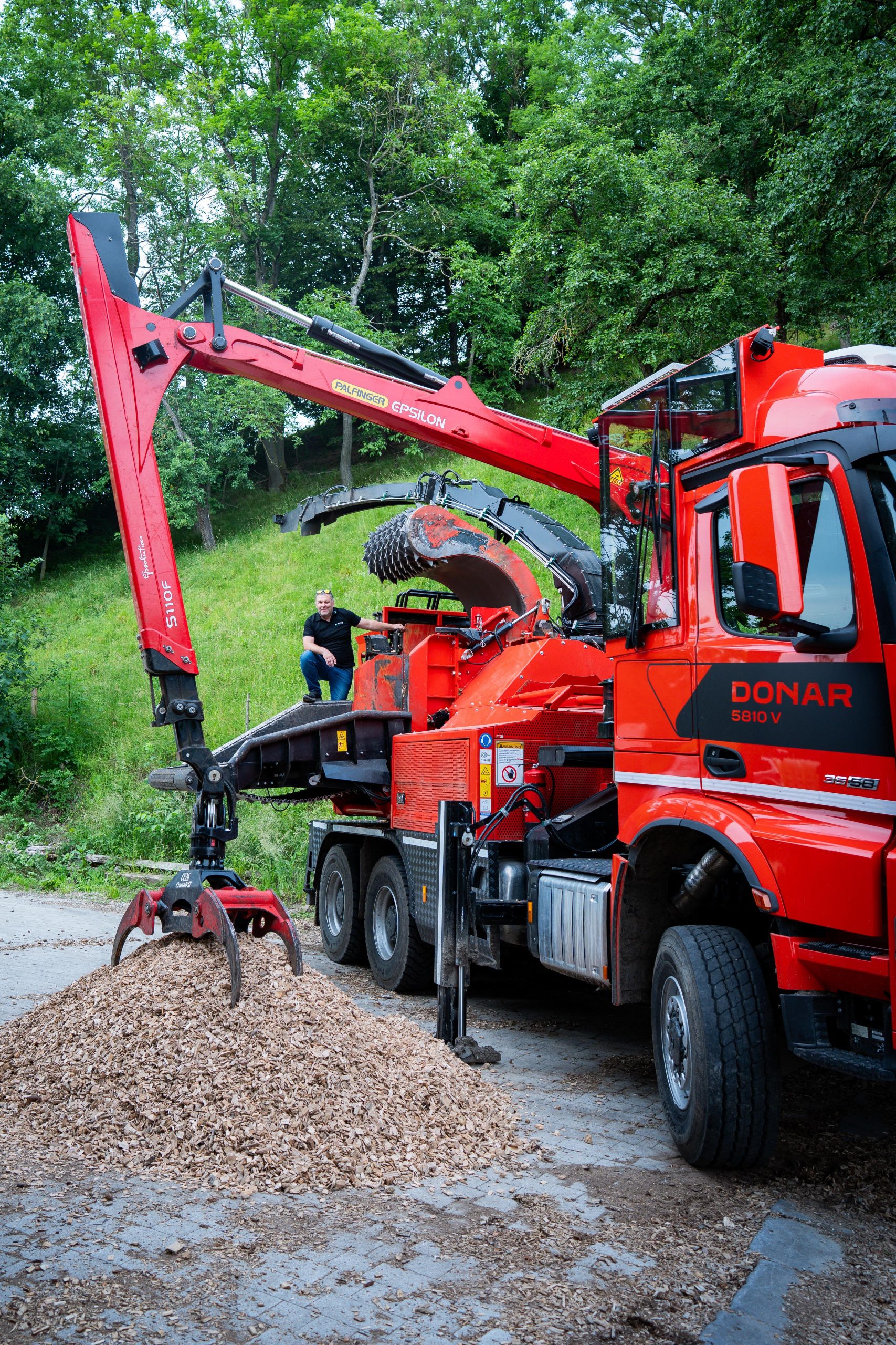 Eine große mobile Holzhackmaschine vom Typ DONAR 5810 V mit rotem PALFINGER EPSILON Ladekran verarbeitet Holz zu Hackschnitzeln. Der Auswurf liegt auf einem Haufen am Boden. Ein Mann in schwarzem Shirt steht auf der Maschine, hinter ihm ein bewaldeter Hang. Das Bild zeigt nachhaltige Technik im Einsatz